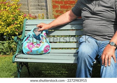 Thief sitting on a park bench next to an unattended bag or purse and stealing property from it. Royalty-Free Stock Photo #2272983915