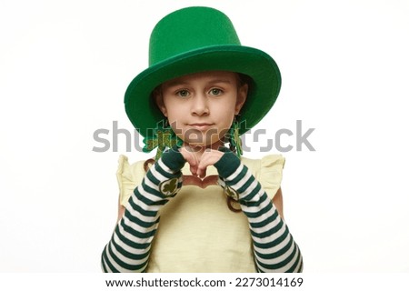 Adorable Irish little child girl, dressed as Leprechaun in green carnival wear for St Patrick's Day, showing a heart shape from fingers, isolated on white background. Ireland's culture and traditions