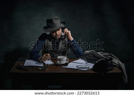 Professional detective in fedora hat sitting at table and talking on phone over dark green vintage background. Investigation. Concept of occupation, character, history. Retro style