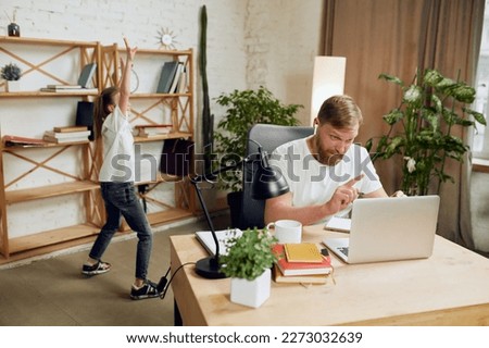 Mature man, father sitting at table, having online video call and working on laptop remotely at home with little girl playing. Concept of fatherhood, childhood, family, freelance job, home office