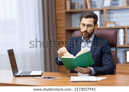 A young man is sitting in the office in a suit at the table, holding and concentrating on reading a green book. Resting, studying, break.