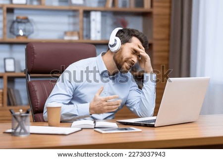 A young male businessman is sitting in the office in headphones and talking on a video call from a laptop. Unpleasant conversation, problems with communication, conference. Worriedly holding his head. Royalty-Free Stock Photo #2273037943