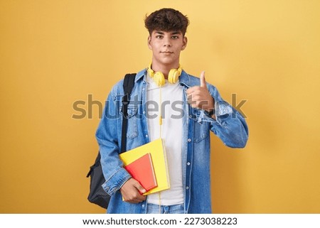 Hispanic teenager wearing student backpack and holding books doing happy thumbs up gesture with hand. approving expression looking at the camera showing success. 