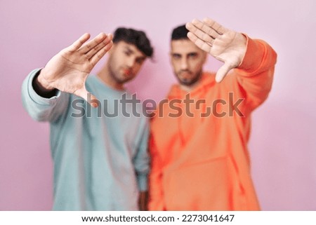 Young hispanic gay couple standing over pink background doing frame using hands palms and fingers, camera perspective 