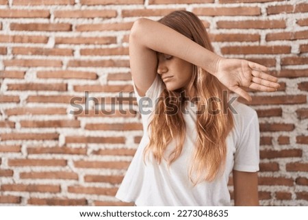 Young caucasian woman standing over bricks wall covering eyes with arm, looking serious and sad. sightless, hiding and rejection concept 