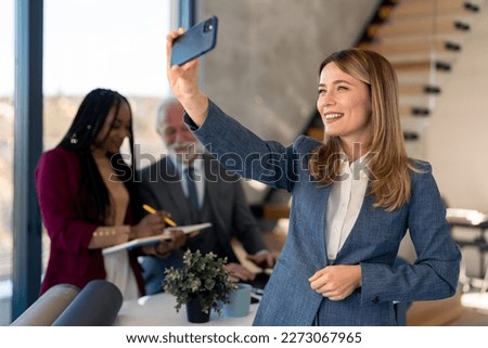 Beautiful millennial businesswoman wearing blue business suit and white shirt using smart mobile phone to make self portrait photo while standing at modern workplace with diverse business coworkers.