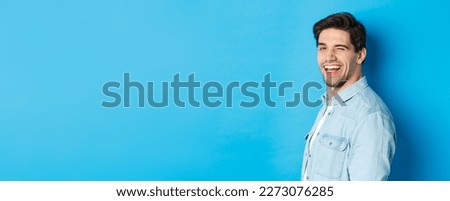 Happy and confident man turn head at camera, winking and smiling, standing over blue background. Royalty-Free Stock Photo #2273076285