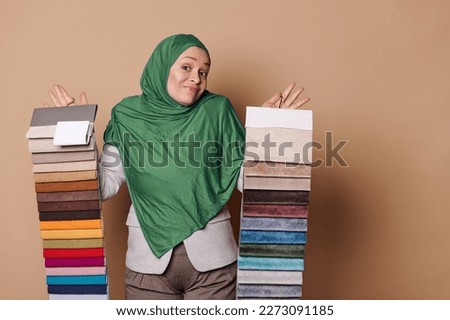 Charming Arab Muslim woman in green hijab, feels doubt while chooses upholstery textile, holds fabric samples on beige isolated background. Interior design. Home improvement, decoration, renovation Royalty-Free Stock Photo #2273091185