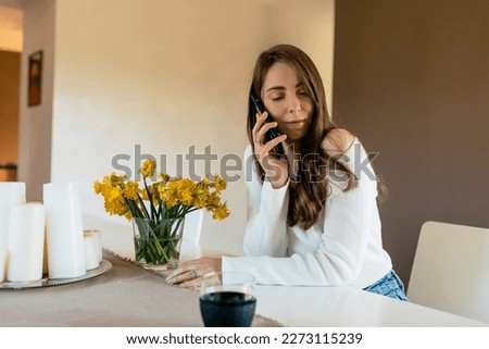 Positive happy caucasian woman in good mood sits at the table near fresh flowers in morning. Brunette with wavy hair wears shirt. People emotions, lifestyle and fashion concept.