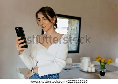 Lovely cute girl with dark collected hair wearing white shirt is using smartphone with happy smile and standing in the living room in sunny warm day. Royalty-Free Stock Photo #2273115265