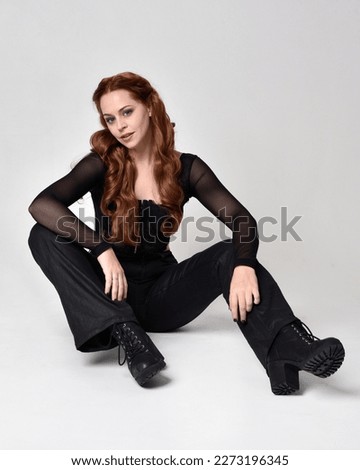 Full length portrait of beautiful woman with long red hair wearing corset top  leather pants.  Sitting pose with gestural hands reaching out, sitting  on floor. Isolated on white studio background