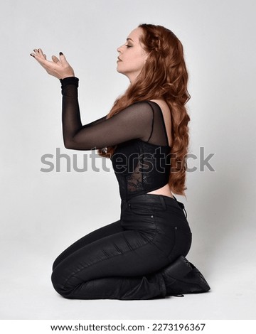 Full length portrait of beautiful woman with long red hair wearing corset top  leather pants.  Sitting pose with gestural hands reaching out, sitting  on floor. Isolated on white studio background