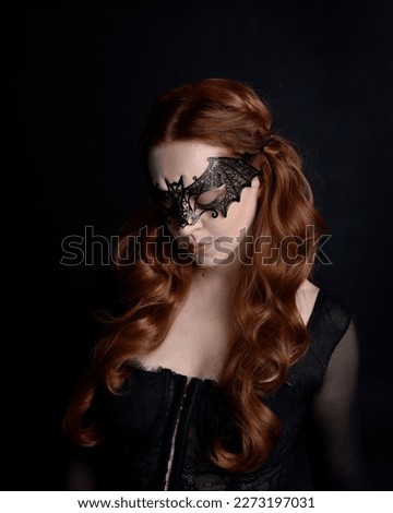 Masked close up portrait of beautiful woman with long red hair wearing sheer corset top, and black bat wing mask.  Isolated on dark studio background with. Moody silhouette lighting.