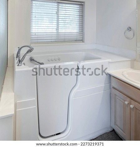 Square Walk-in bathtub with elderly and handicapped accessibility. There is a vanity sink with mirrors on the right and shower stall with frosted glass and aluminum frames. Royalty-Free Stock Photo #2273219371