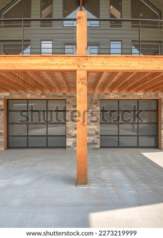 Vertical Two glass garage door of a modern custom house with stone veneer siding. Garage driveway wooden deck with metal railing against the gray siding and picture windows at the second floor.