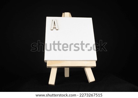 A wooden small capital letter and white blank painting canvas resting on a miniature artists easel isolated on a black background