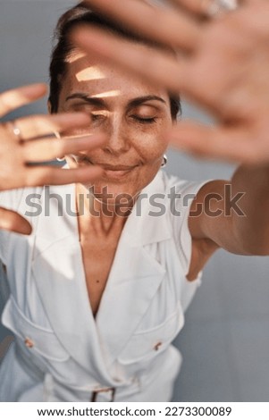 Close up portrait beauty brunette middle age woman alone in white clothes standing in sunlight and covering face with hand from bright light in summer day outdoor. Summer time