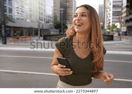 Laughing girl holding mobile phone looking to the side in empty metropolis avenue Royalty-Free Stock Photo #2273344897