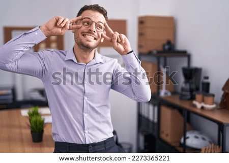 Young hispanic man at the office doing peace symbol with fingers over face, smiling cheerful showing victory 