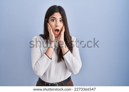 Young brunette woman standing over blue background afraid and shocked, surprise and amazed expression with hands on face 