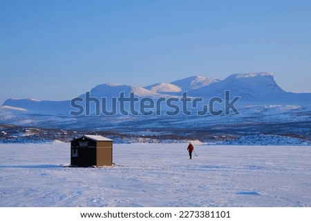 Wooden house standing on skids on frozen lake Torneträsk (Tornestrask) around Abisko National Park. Silhouette of person on snowshoes on lake. Sweden, Arctic Circle, Swedish Lapland