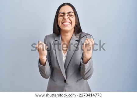 Hispanic business woman wearing glasses very happy and excited doing winner gesture with arms raised, smiling and screaming for success. celebration concept.  Royalty-Free Stock Photo #2273414773