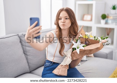 Caucasian woman holding bouquet of white flowers taking a selfie picture puffing cheeks with funny face. mouth inflated with air, catching air. 