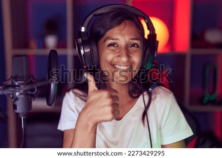 Young hispanic woman playing video games wearing headphones smiling happy and positive, thumb up doing excellent and approval sign 