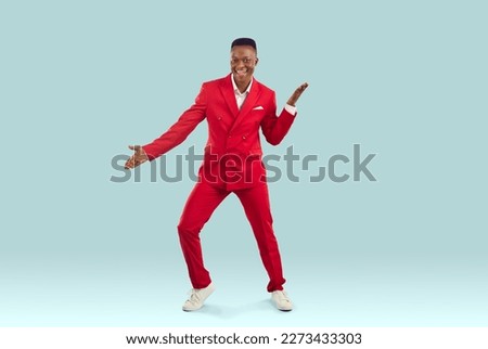 Happy handsome African American guy in stylish red suit dancing in fashion studio. Joyful attractive Tanzanian man in funky jacket and trousers dancing to lively music on light blue colour background