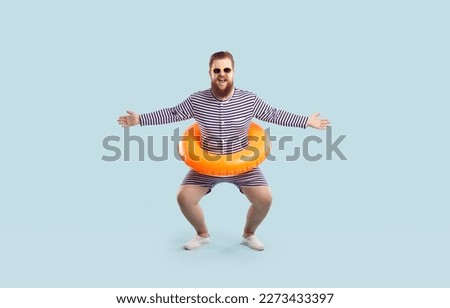 Funny fat man in retro swimsuit and sunglasses poses with swim tube around his waist isolated on blue background. Happy plump bearded man with swim ring having fun and enjoying summer holiday at beach Royalty-Free Stock Photo #2273433397
