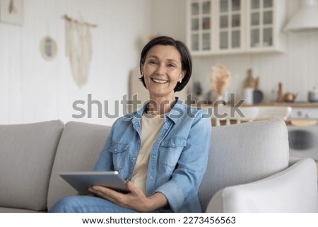 Cheerful attractive senior freelance woman using gadget at home, holding tablet computer, enjoying wireless domestic Internet connection, online app, web service, smiling at camera. Female portrait Royalty-Free Stock Photo #2273456563