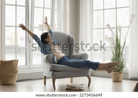 Excited carefree mature lady enjoying being in cozy stylish home interior, leisure, relaxation, resting in soft armchair, stretching body, rising hands in joy, celebrating, feeling happy