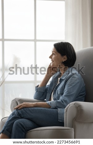 Happy dreamy pretty mature woman relaxing in armchair at home, thinking on good future plans, touching chin, looking away, smiling, dreaming. Positive senior lady enjoying leisure at home