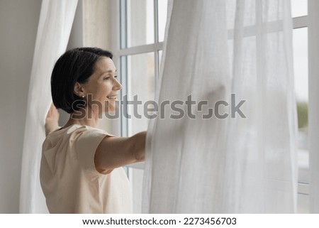 Happy mature homeowner woman looking out of big window, enjoying view, daylight, good morning, parting white transparent veils in bedroom, opening drapes, curtains