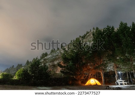 A tourist tent and a table with a lantern glowing at night in the dark against the background of trees and mountains with a sky and a fence.