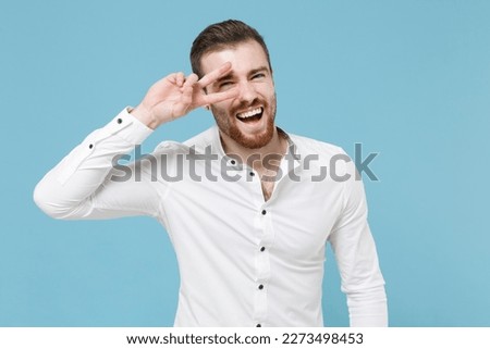 Cheerful young bearded man guy 20s in white classic shirt isolated on pastel blue wall background studio portrait. People sincere emotions lifestyle concept. Mock up copy space. Showing victory sign