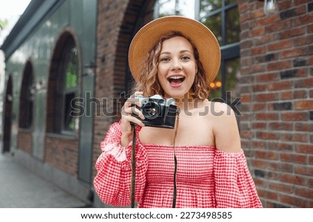 Young surprised happy excited woman wear pink dress hold take picture on retro vintage photo camera walk in city standing outdoor near town brick building. People urban summer time lifestyle concept.