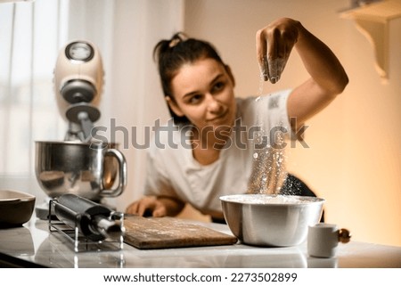 Selective focus on hand of woman pouring white flour into stainless bowl on table with modern kitchen appliances. Woman prepares dough pouring flour. Royalty-Free Stock Photo #2273502899