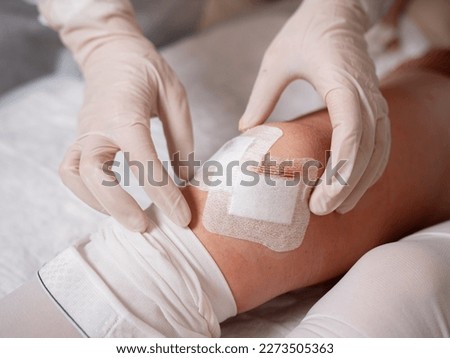 The nurse's hands in medical gloves stick a sterile patch on the incision in the patient's knee after arthroscopy surgery.