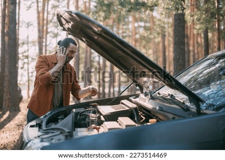 A young upset man calls by phone to a car service near a broken car far outside the city. The car broke down on a trip. Calling a tow truck. Royalty-Free Stock Photo #2273514469