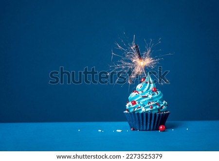Blue cupcake with red and white sprinkles and lit sparkler on a blue background.