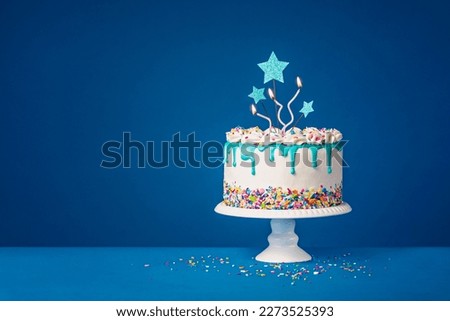 White birthday drip cake with teal ganache, star toppers and fun candles over dark blue background