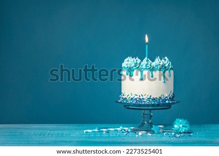 White birthday drip cake with teal ganache and lit candle over blue background
