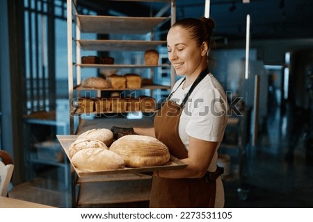 Pretty woman baker holding variety of baked bread on tray Royalty-Free Stock Photo #2273531105