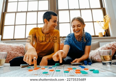 Two young women sitting on couch playing board game with counters, laughing. Happy couple bonding over an indoor leisure activity, smiling and sitting together at a table. Royalty-Free Stock Photo #2273535689