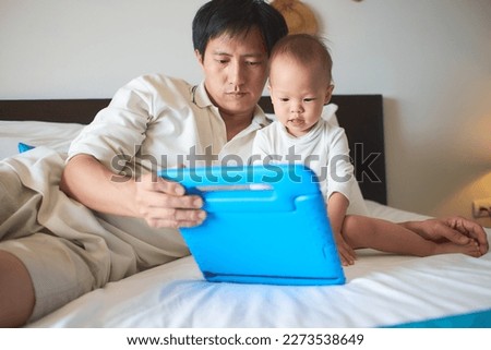 Father and son are using tablet while spending time together before go to bed at home, Dad and Cute little Asian 1 year old toddler baby boy child sitting on bed in bedroom, internet addiction concept