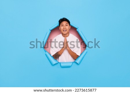 Happy Asian man put palms together gesturing a pray sign, he is asking for something with positive expression, isolated next to torn blue wall