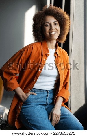 Authentic portrait of beautiful smiling African American woman wearing stylish casual orange t shirt, jeans sitting at home. Happy confident curly haired hipster female looking at camera 