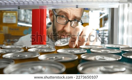 Close-up of the hand of a bearded man with glasses taking one can of soda or energy drink in a store fridge