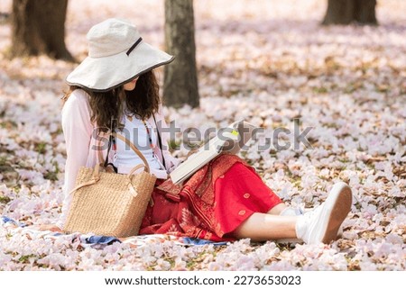 young girl long leg read book sitting on Falling pink flowers over ground. Woman enjoy read book in park at autumn many pink flower falling like flower courtyard